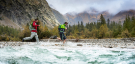two hikers, in a mountain valley, running through a river/stream to get to the other side. Misty mountains in the background. Ross Woodhall - Action & Lifestyle Photography