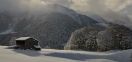 Little Wooden hut covered in snow, surrounded by snow with snow laden trees on the mountain in the distance & also by the hut. Ross Woodhall - Action & Lifestyle Photography