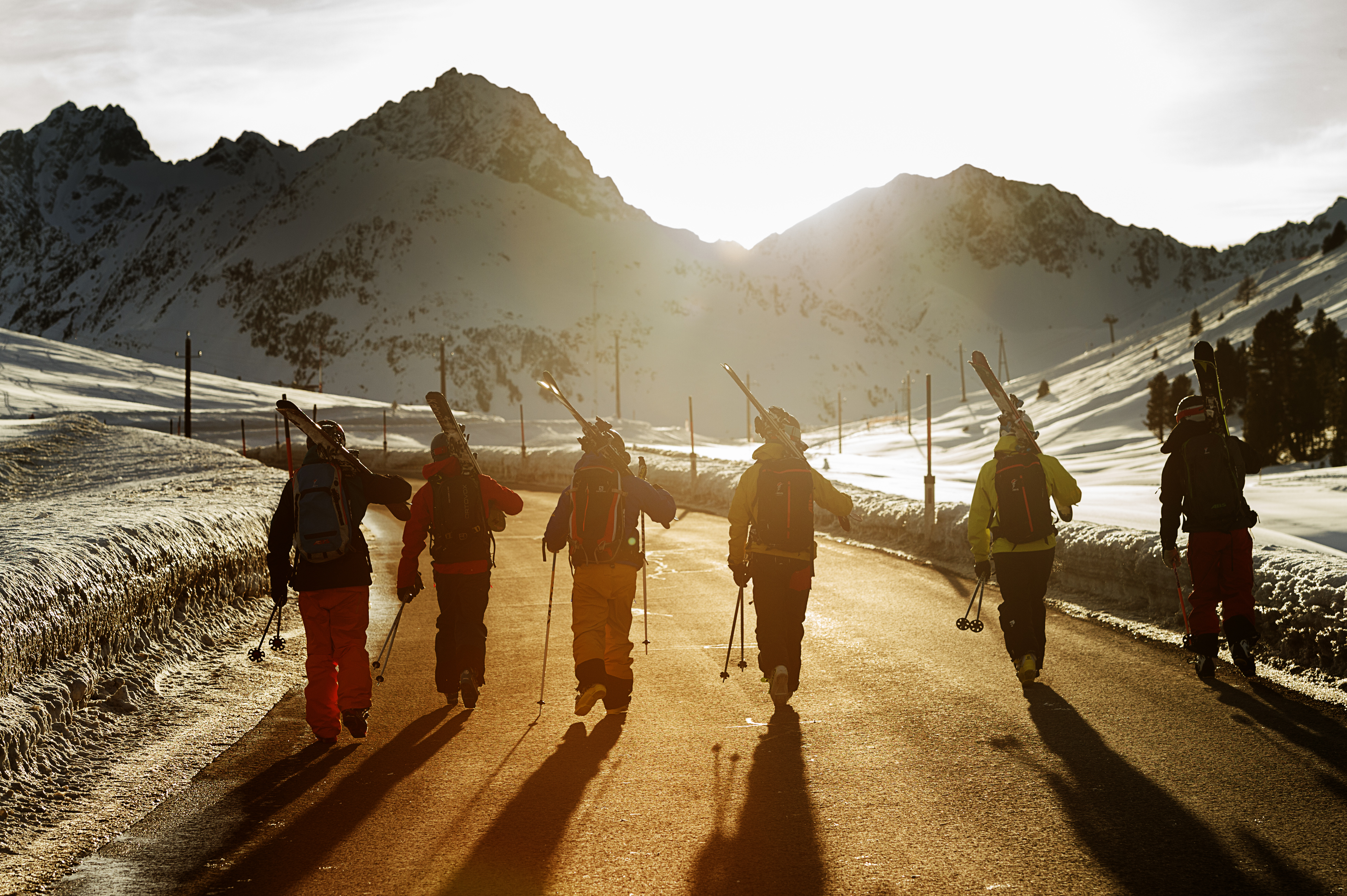 6 skiers walking away from the camera on a road in a alpine area with skis over their shoulders Ross Woodhall - Action & Lifestyle Photography