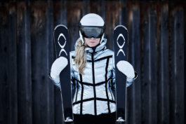 Female skier posing with skis in each hand in front of a wooden hut, wearing a silver jacket with black line details, also wearing a helmet, googles & mittens. Ross Woodhall - Action & Lifestyle Photography